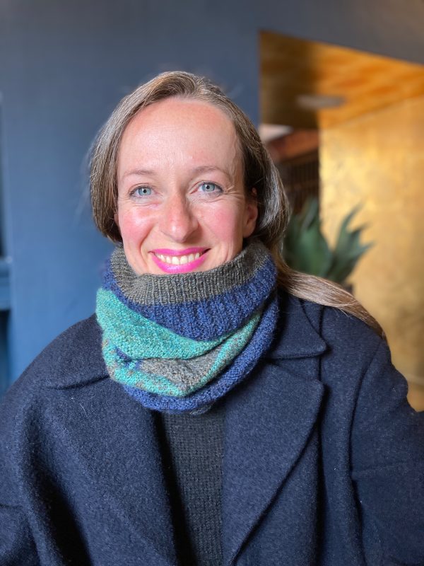 A smiling person is wearing a navy, green and brown knitted cowl